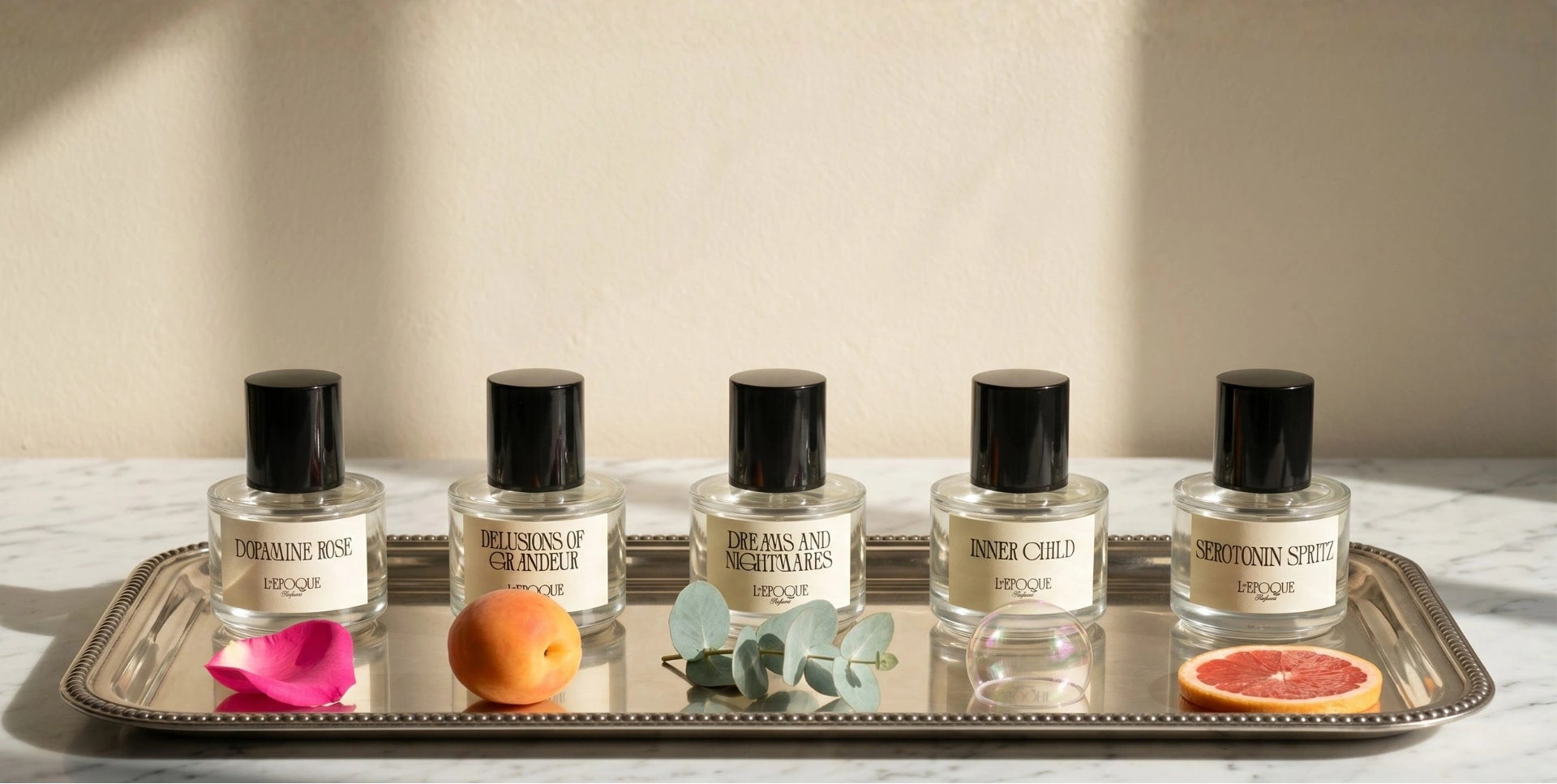 L'Epoque Parfums full collection of five 30mL fragrances on a silver tray: Dopamine Rose extrait de parfum, Delusions of Grandeur eau de parfum, Dreams and Nightmares eau de parfum, Inner Child eau de parfum, and Serotonin Spritz eau de parfum, styled with a rose petal, apricot, eucalyptus sprig, soap bubble, and grapefruit slice