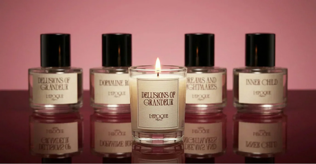 L'Epoque Parfums Delusions of Grandeur mini candle with lit flame, surrounded by the full fragrance collection on a pink background