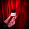 Hand holding a small perfume bottle labeled 'L'Epoque' against a red curtain background