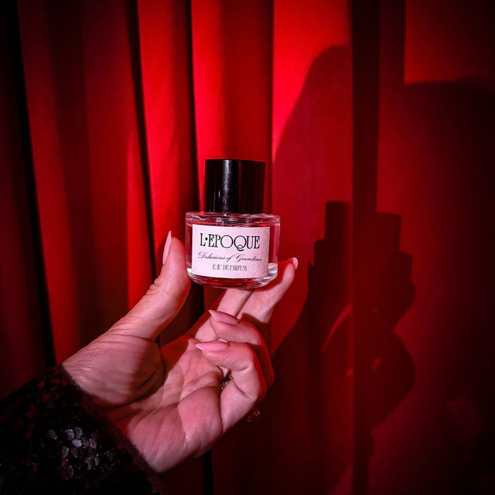Hand holding a small perfume bottle labeled 'L'Epoque' against a red curtain background