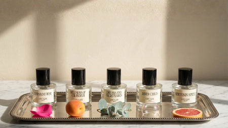 L'Epoque Parfums full collection of five 30mL fragrances on a silver tray: Dopamine Rose extrait de parfum, Delusions of Grandeur eau de parfum, Dreams and Nightmares eau de parfum, Inner Child eau de parfum, and Serotonin Spritz eau de parfum, styled with a rose petal, apricot, eucalyptus sprig, soap bubble, and grapefruit slice