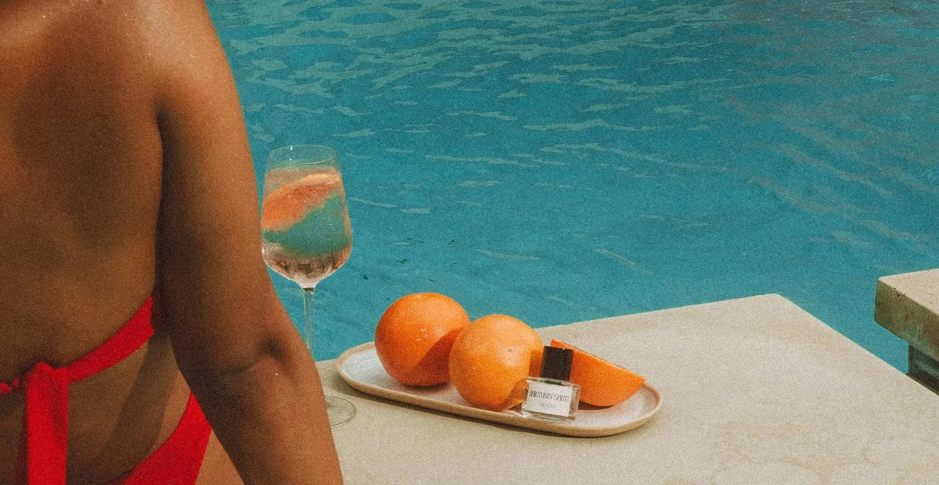 Serotonin Spritz Eau de Parfum by L'Epoque on a ceramic tray with grapefruit and a spritz cocktail at the sunlit edge of a pool, a hand resting on the warm stone beside it