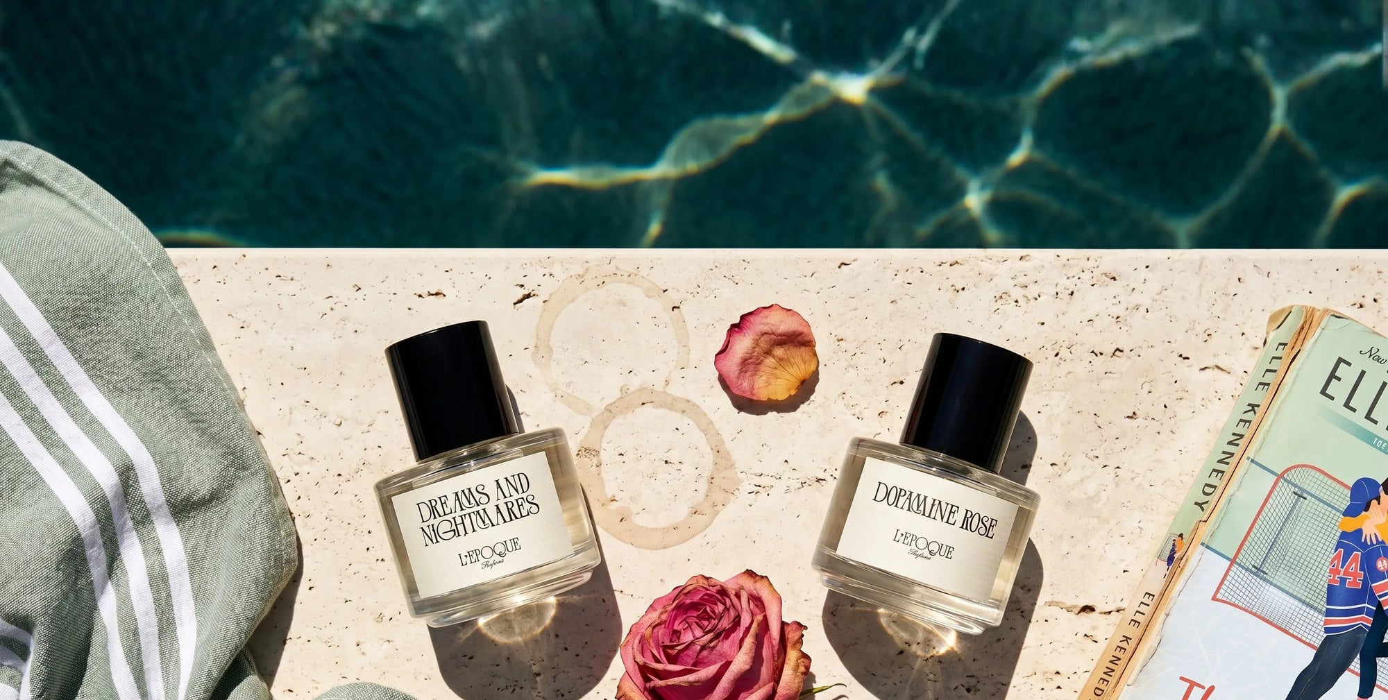 Dreams and Nightmares and Dopamine Rose perfumes by L'Epoque Parfums resting poolside on travertine, surrounded by a dried rose, rose petals, and a well-loved paperback