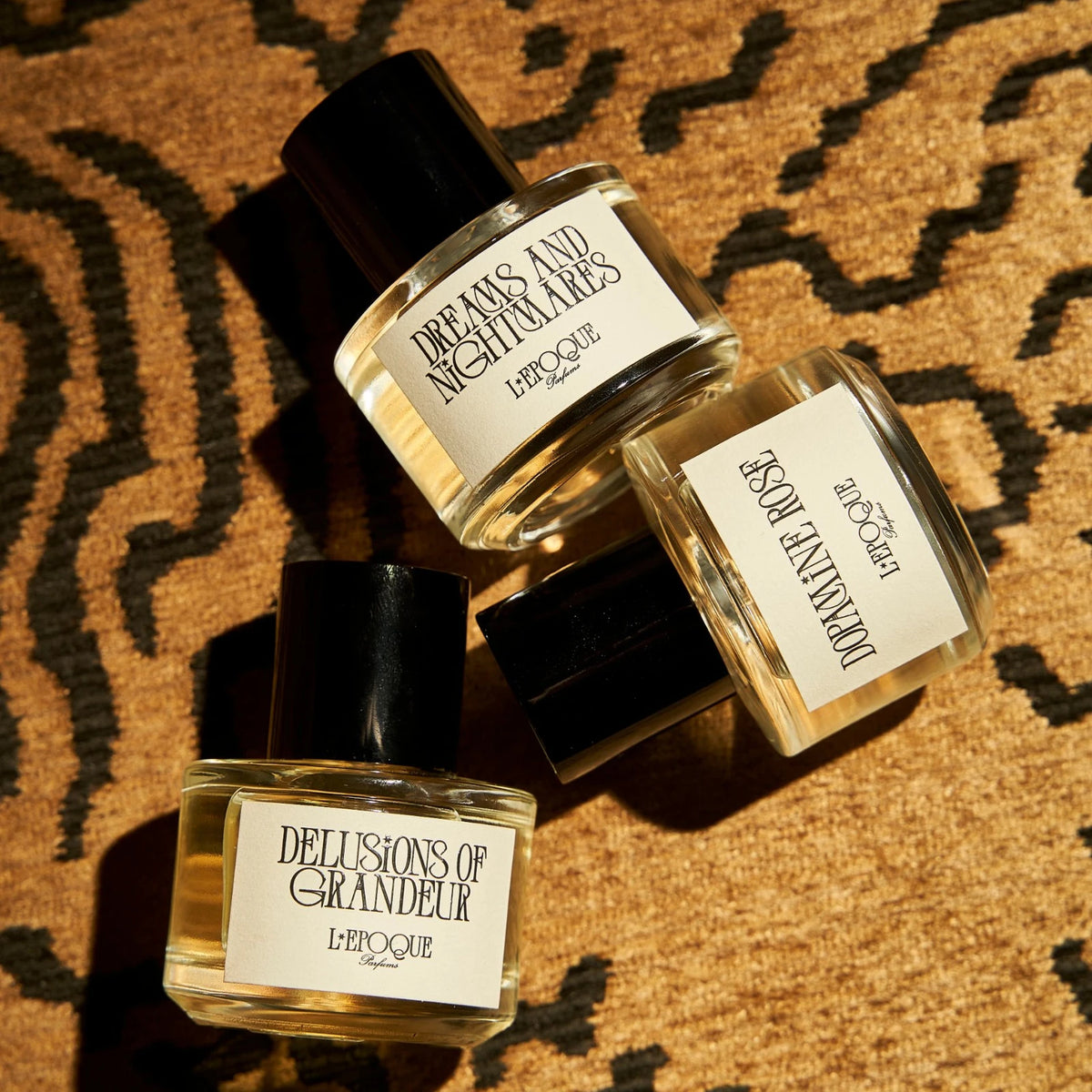 Delusions of Grandeur eau de parfum 30mL by L’Epoque, held up in a man’s hand beside a woman in a white top with red nails, cap in hand